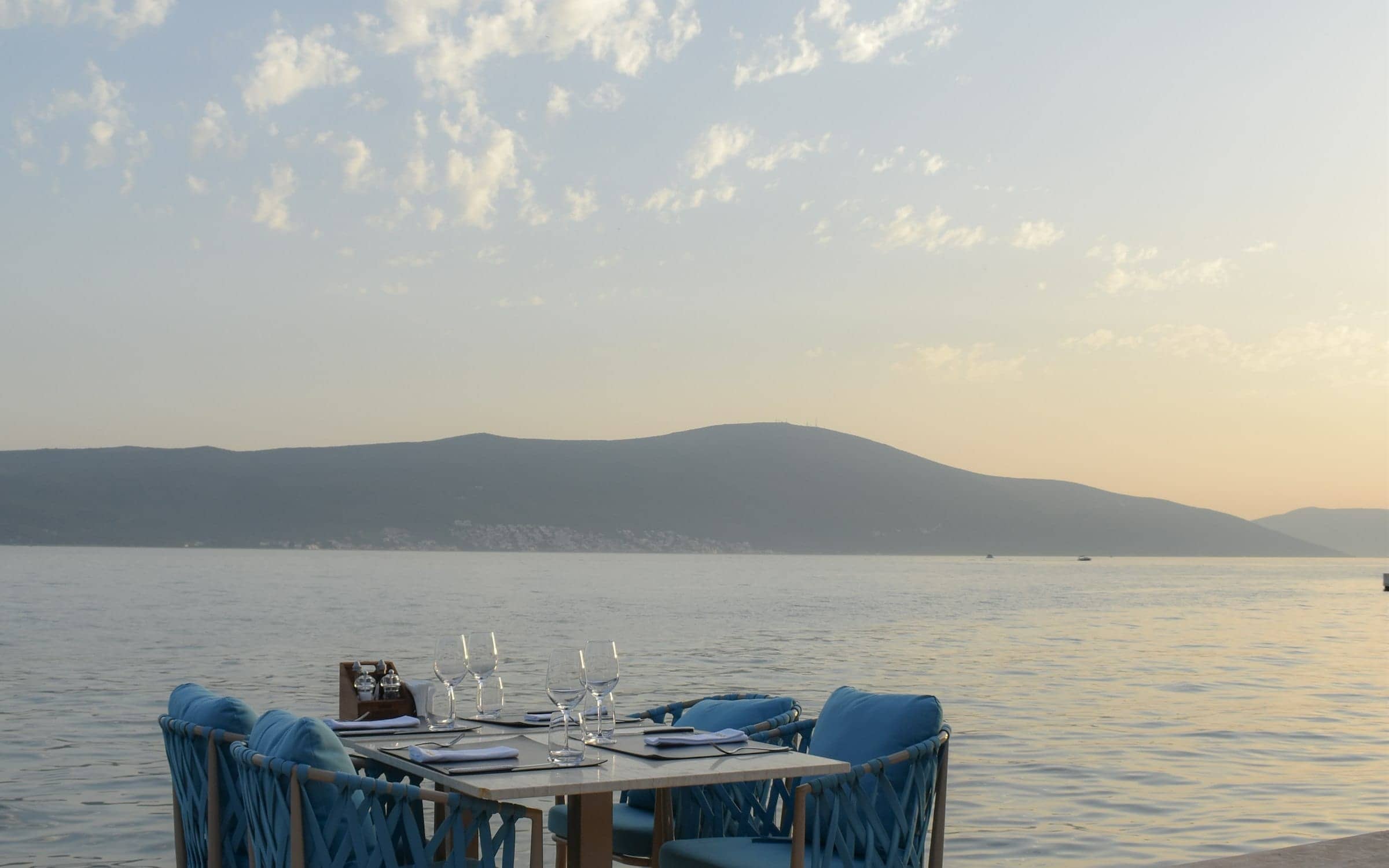 A table laid with glasses at the water’s edge, mountains beyond at dusk